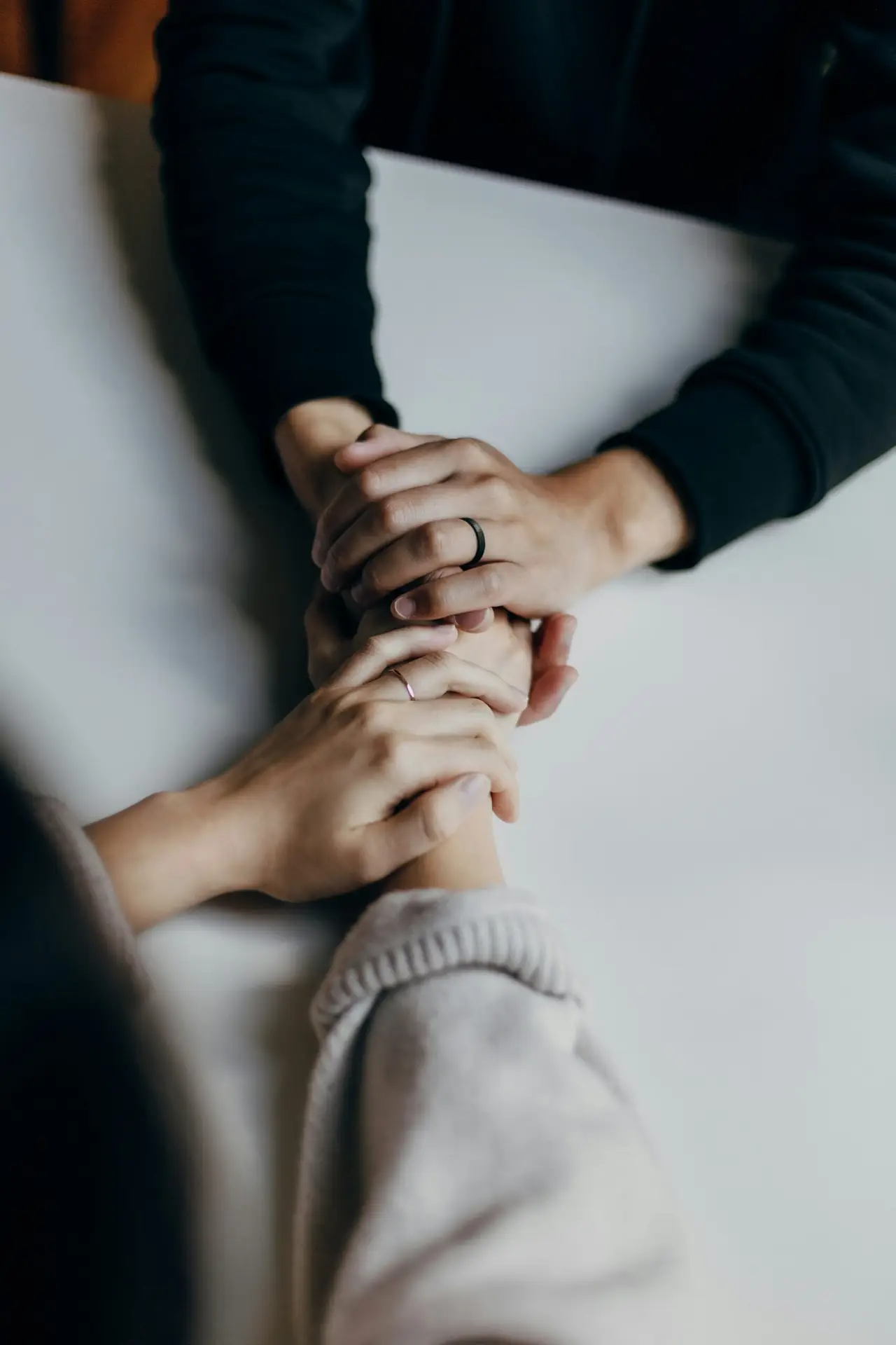 Two people sit across from each other at a table, gently holding hands. One person is wearing a dark long-sleeve top and a ring, while the other person is wearing a light-colored sleeve.
