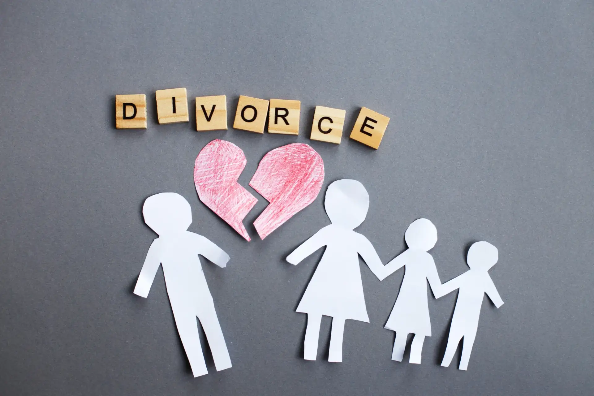 Paper cutouts of a man, woman, and two children stand under a broken red heart and wooden letter tiles spelling “DIVORCE,” symbolizing the creation of an Alternate Home on a gray background.
