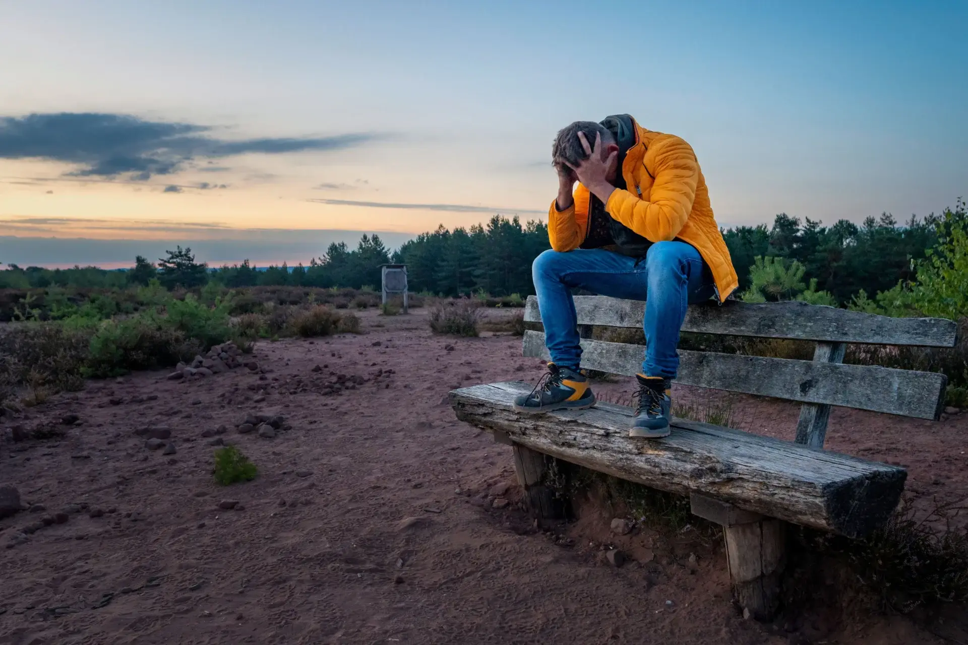 A person in a yellow jacket sits on a wooden bench outdoors, hunched over with their head in their hands, appearing distressed. The background features trees, shrubs, and a sunset sky.